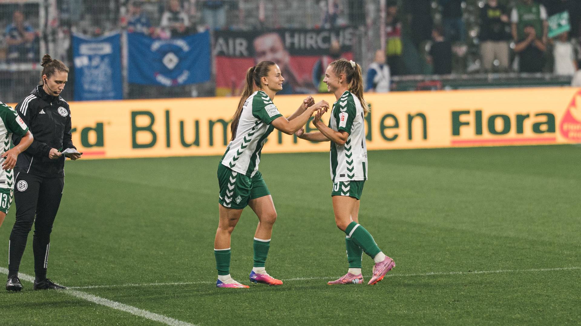 Tuana Mahmoud und Michelle Weiß, die grüne Trikots tragen, geben sich auf dem Spielfeld die Hand, während ein Trainer daneben steht und im Hintergrund die Zuschauer zu sehen sind.