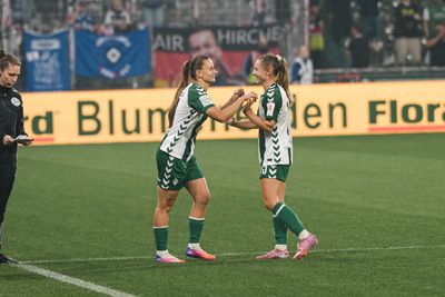 Tuana Mahmoud und Michelle Weiß, die grüne Trikots tragen, geben sich auf dem Spielfeld die Hand, während ein Trainer daneben steht und im Hintergrund die Zuschauer zu sehen sind.
