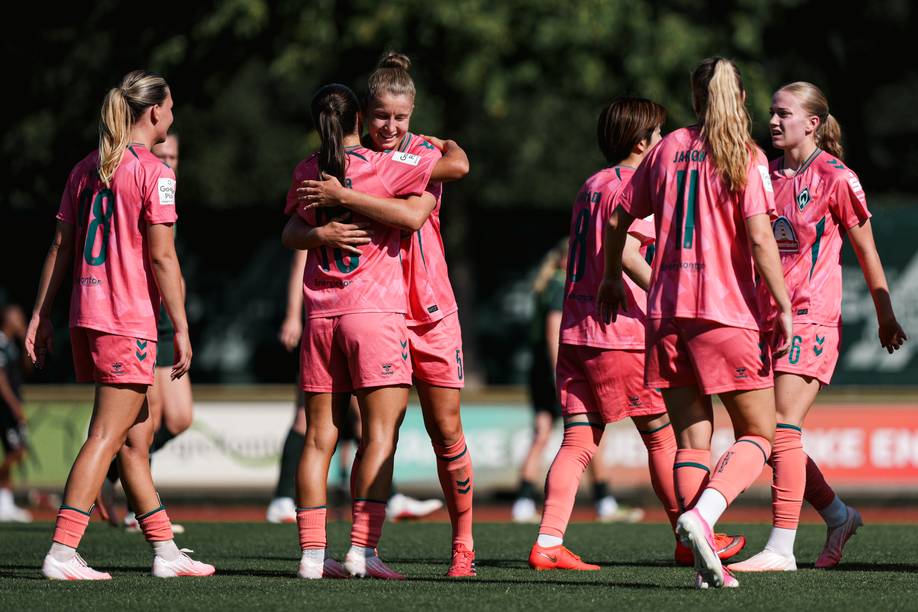 Eine Frauenfußballmannschaft in rosa Trikots feiert auf dem Spielfeld; zwei Spielerinnen umarmen sich, während andere unter strahlendem Sonnenschein daneben stehen.
