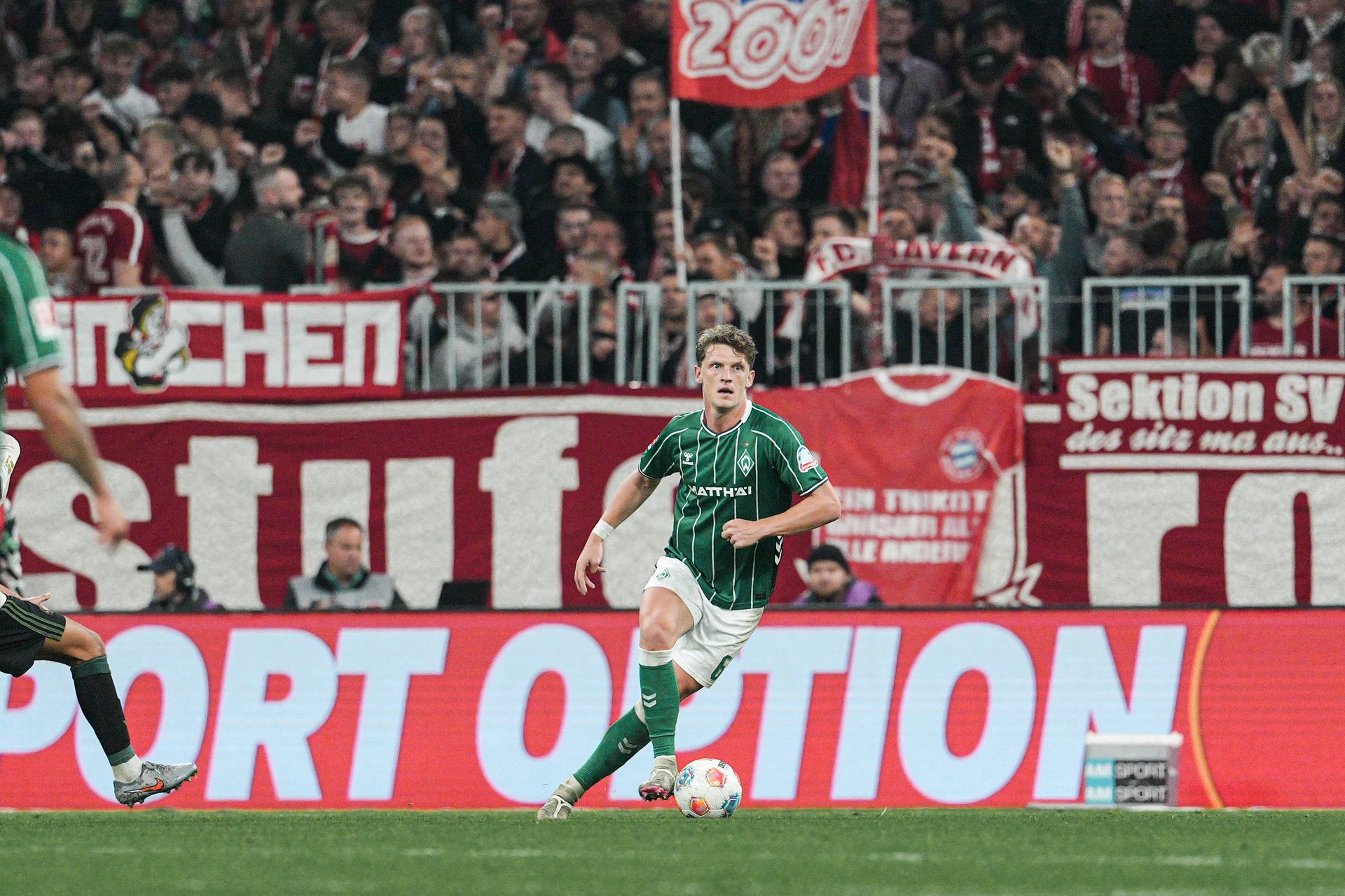 Jens Stage mit Ball am Fuß, im Hintergrund die Bayern-Kurve.