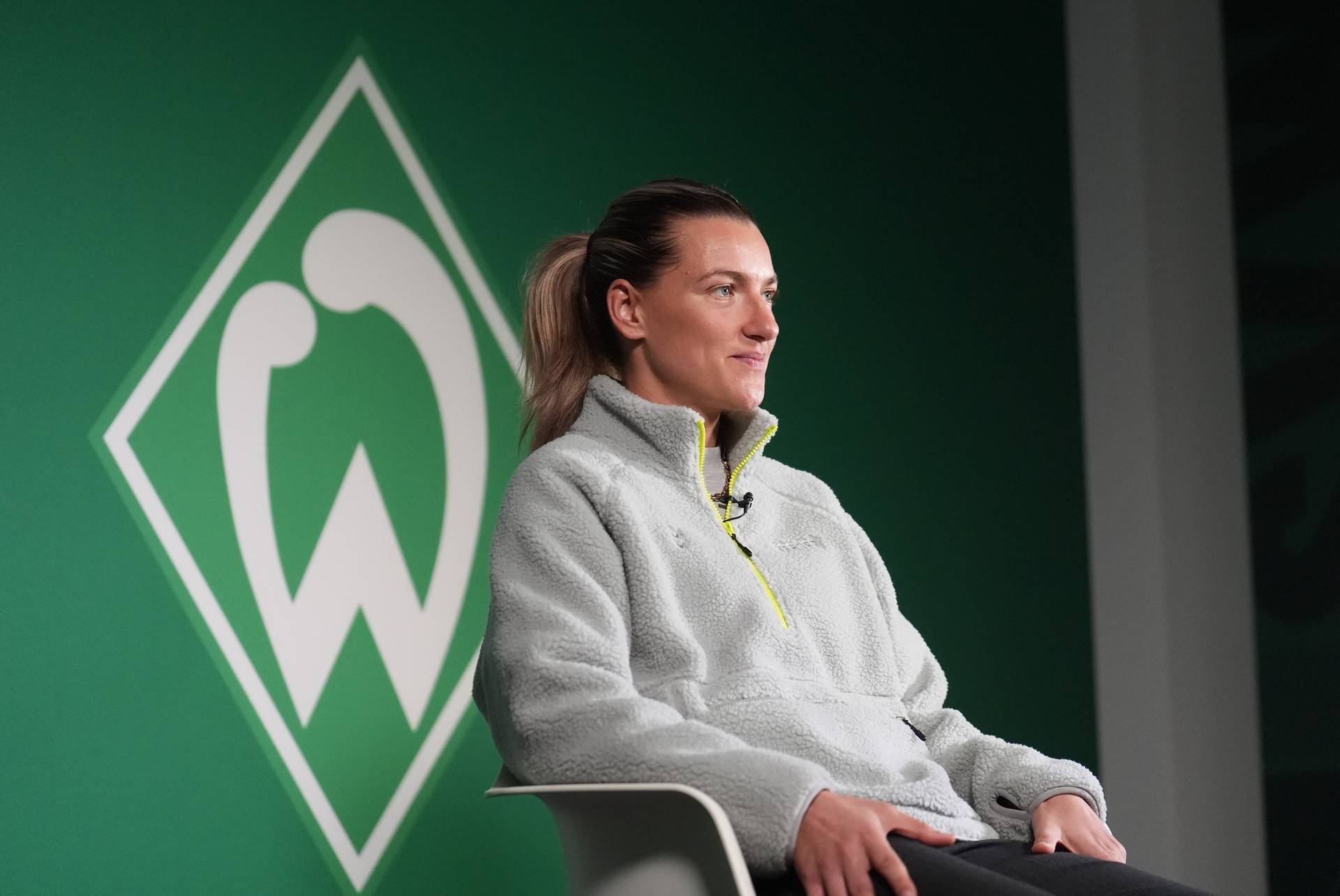Medina Desic sitzt vor dem Werder-Logo auf einem Stuhl und lächelt.
