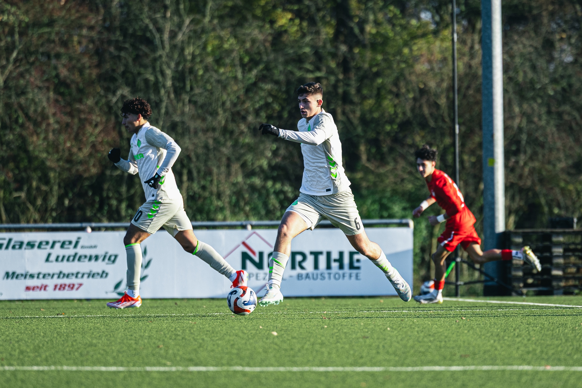Zwei Fußballspieler in weißen Trikots sprinten über einen Rasen, im Hintergrund ist ein weiterer Spieler in Rot zu sehen, hinter ihm sind Werbeplakate zu erkennen.