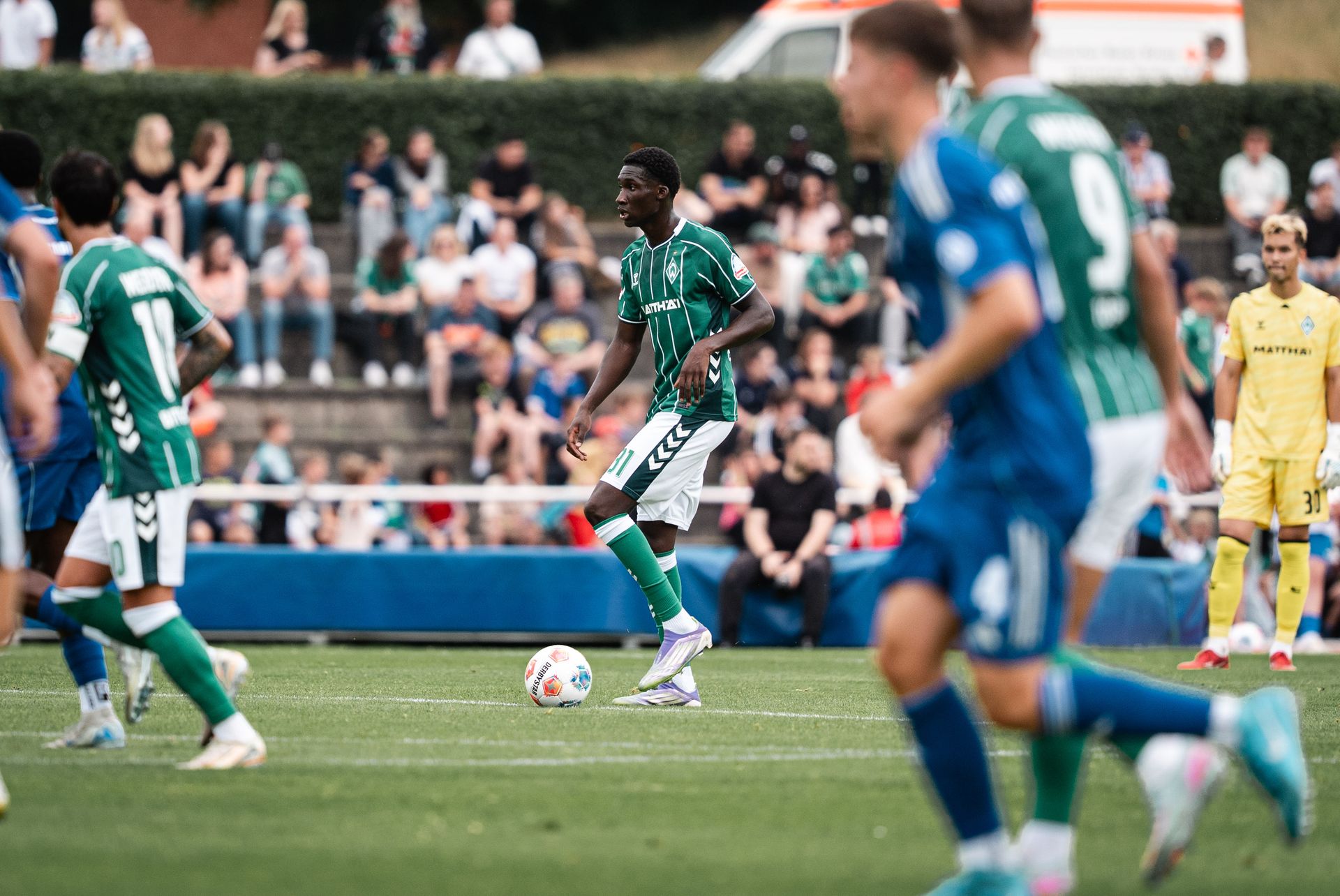 Soccer players in green and blue jerseys on a field, with one player dribbling the ball, and a crowd in the background.
