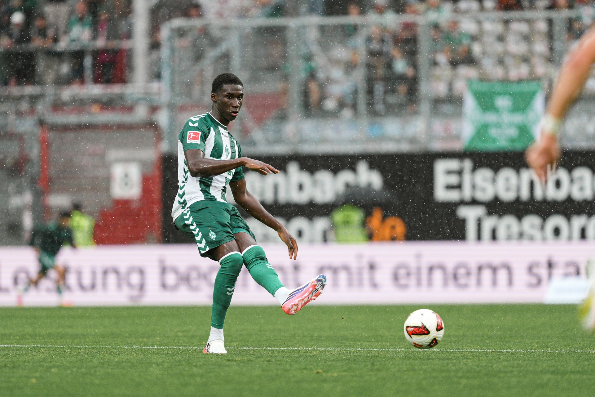 Soccer player in green and white uniform about to kick a ball on a wet field, with spectators in the background.
