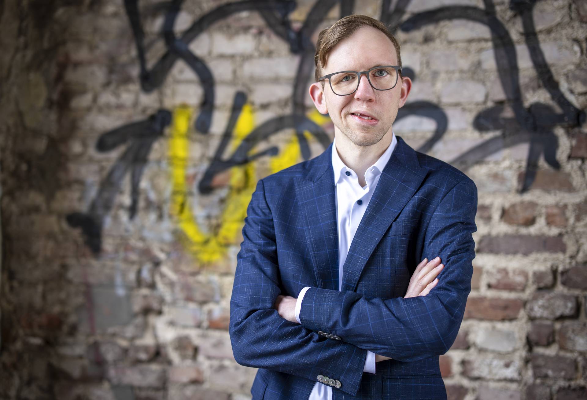 Ein Mann mit Brille und blauem Anzug steht mit verschränkten Armen lächelnd vor einer mit Graffiti übersäten Backsteinmauer.