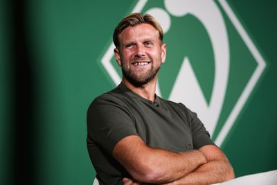 Lächelnder Mann mit kurzen blonden Haaren und Bart, der ein dunkles T-Shirt trägt, die Arme verschränkt, vor einem grünen Hintergrund mit einem Logo.