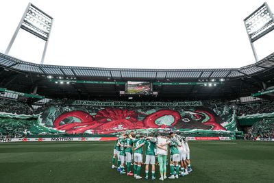 Die Fußballmannschaft versammelt sich auf dem Spielfeld, während auf den Tribünen des Stadions, das voller jubelnder Fans ist, ein großes, farbenfrohes Oktopus-Transparent weht.