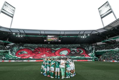Die Fußballmannschaft versammelt sich auf dem Spielfeld, während auf den Tribünen des Stadions, das voller jubelnder Fans ist, ein großes, farbenfrohes Oktopus-Transparent weht.