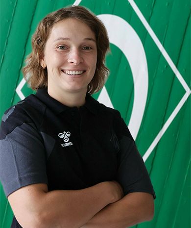 Porträt von Sr. Constanze Hungar. Sie steht mit verschränkten Armen vor einer grünen Wand, auf der die Werder-Raute zu sehen ist.