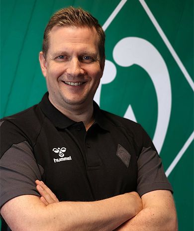 Porträt von Philipp Mehrtens. Er steht mit verschränkten Armen vor einer grünen Wand, auf der die Werder-Raute zu sehen ist.