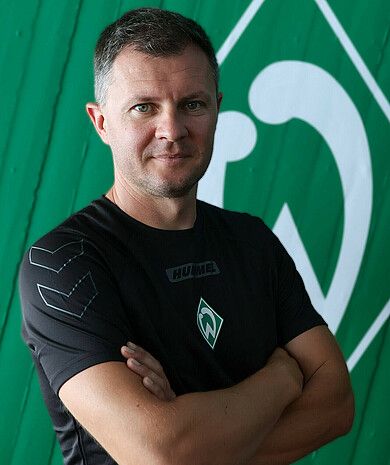 Porträt von Andrei Fabrizius. Er steht mit verschränkten Armen vor einer grünen Wand, auf der die Werder-Raute zu sehen ist.