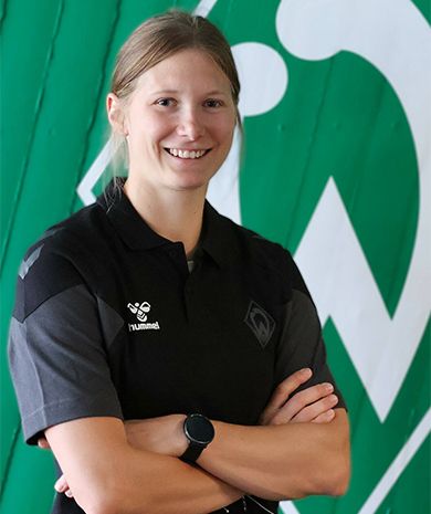 Porträt von Sandra Dinkeldein. Sie steht mit verschränkten Armen vor einer grünen Wand, auf der die Werder-Raute zu sehen ist.