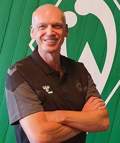 Porträt von Axel Mindermann. Er steht mit verschränkten Armen vor einer grünen Wand, auf der die Werder-Raute zu sehen ist.