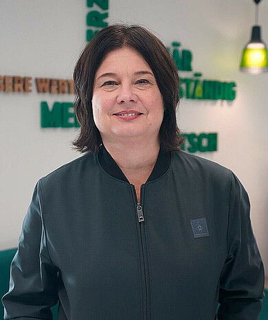 Frau in einer dunklen Jacke lächelt, steht in einem Innenraum mit grüner und brauner Wanddekoration im Hintergrund.