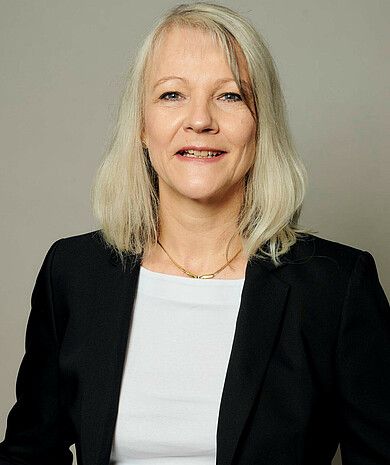 Frau mit schulterlangen blonden Haaren in einem schwarzen Blazer und einem weißen Oberteil, lächelnd vor einem neutralen Hintergrund.