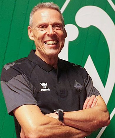 Porträt von Folko Fichtner. Er steht mit verschränkten Armen vor einer grünen Wand, auf der die Werder-Raute zu sehen ist.