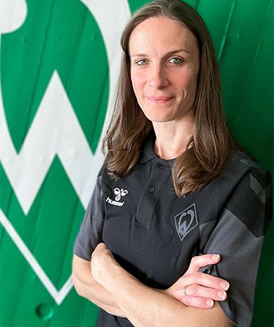 Porträt von Anja Fabrizius. Sie steht mit verschränkten Armen vor einer grünen Wand, auf der die Werder-Raute zu sehen ist.