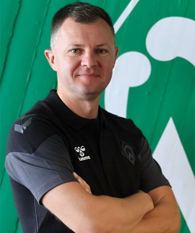 Porträt von Andrei Fabrizius. Er steht mit verschränkten Armen vor einer grünen Wand, auf der die Werder-Raute zu sehen ist.