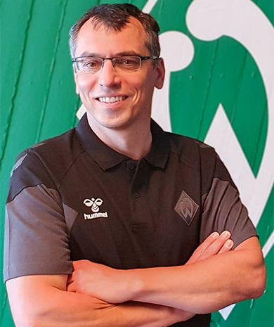 Porträt von Enrico Oelgardt. Er steht mit verschränkten Armen vor einer grünen Wand, auf der die Werder-Raute zu sehen ist.