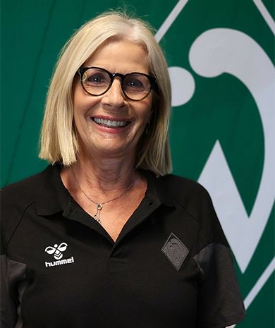 Portrait von Dagmar Stelberg. Sie steht vor einer grünen Wand, auf der die Werder-Raute zu sehen ist.