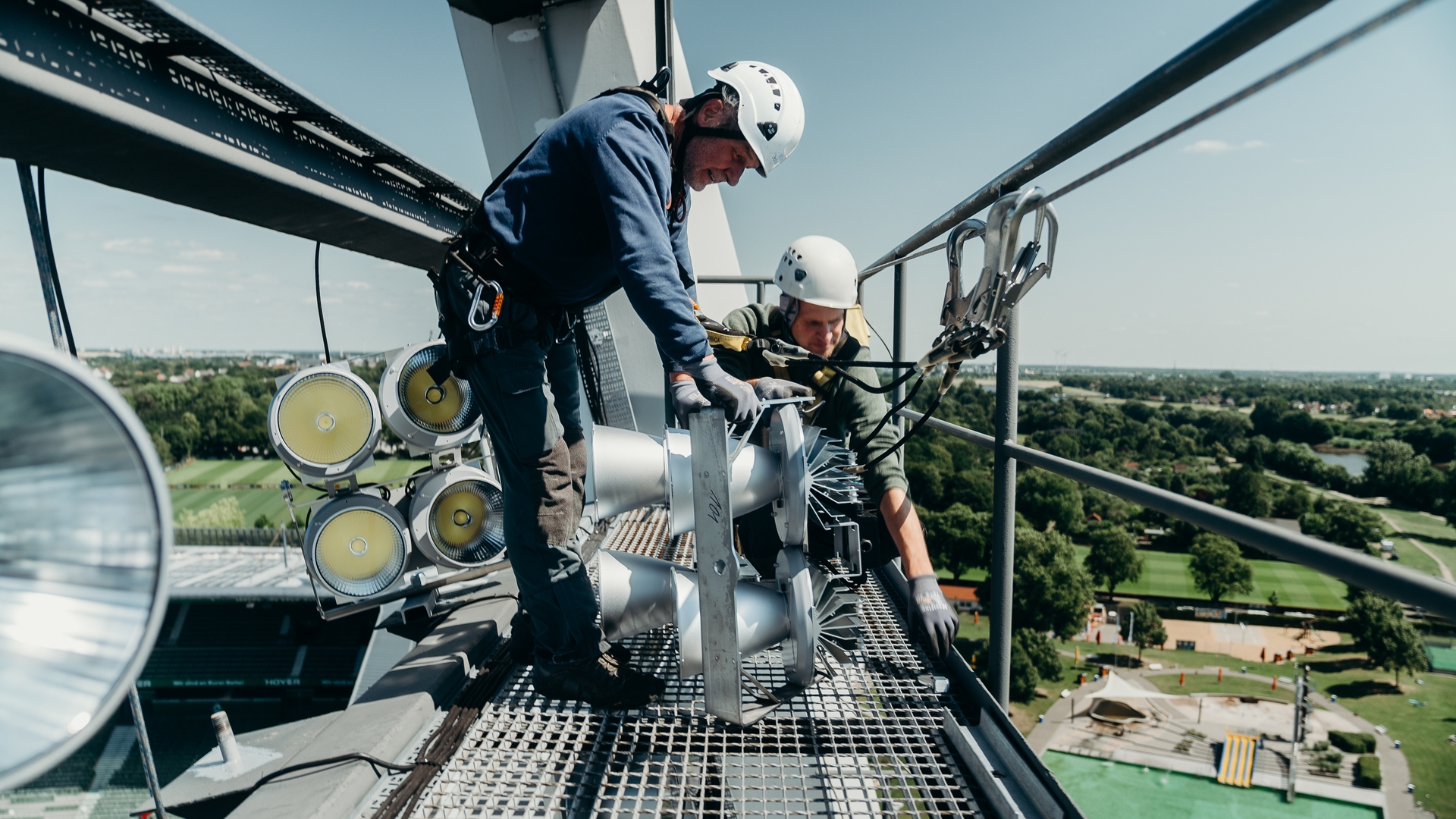 Zwei Arbeiter in Sicherheitsausrüstung installieren Geräte auf einem hohen Bauwerk, im Hintergrund sind die Stadtlandschaft und eine grüne Landschaft zu sehen.