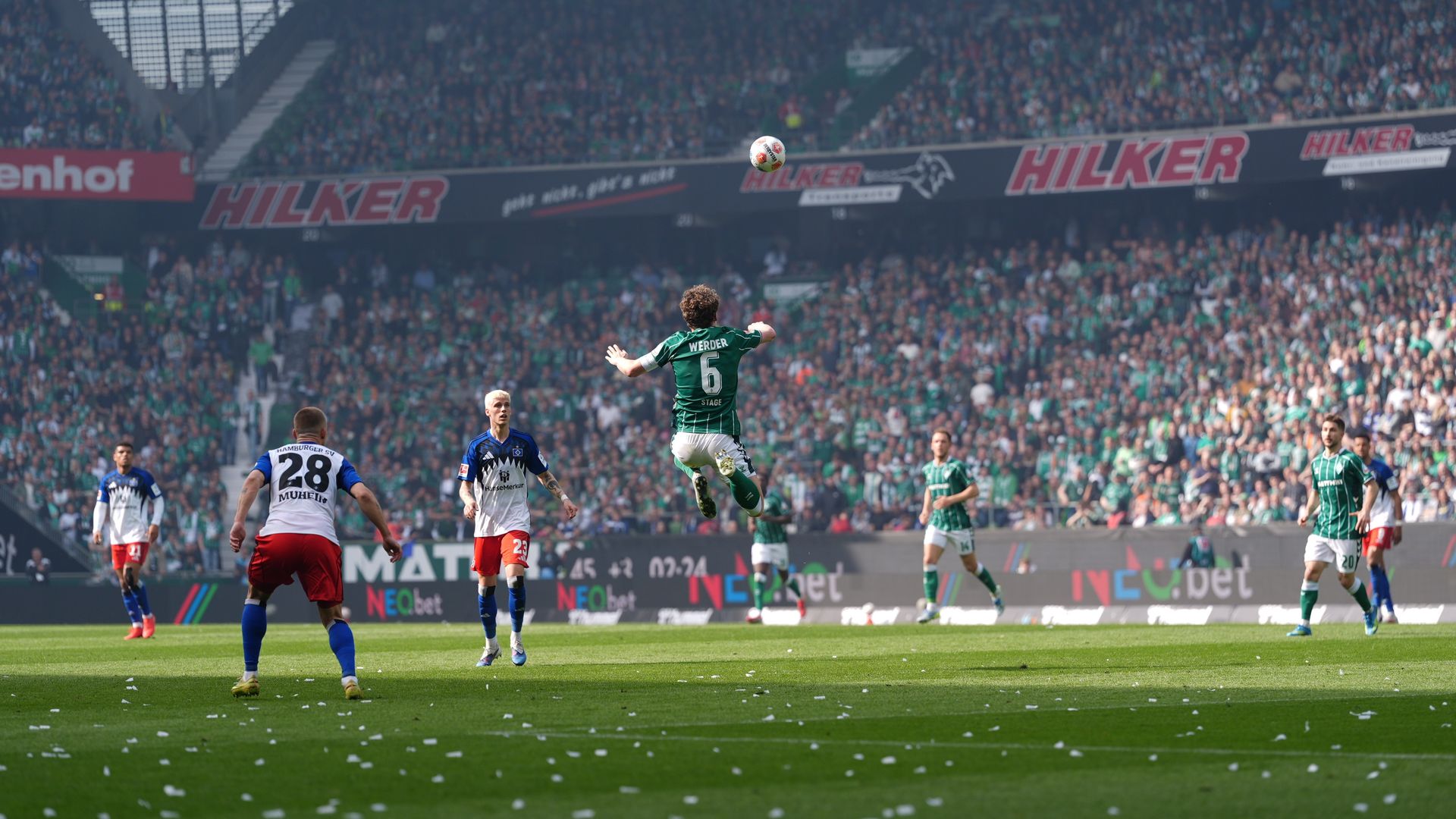 Ein Fußballspieler in grüner Kleidung springt während eines Spiels nach dem Ball, im Hintergrund ist ein vollbesetztes Stadion zu sehen.