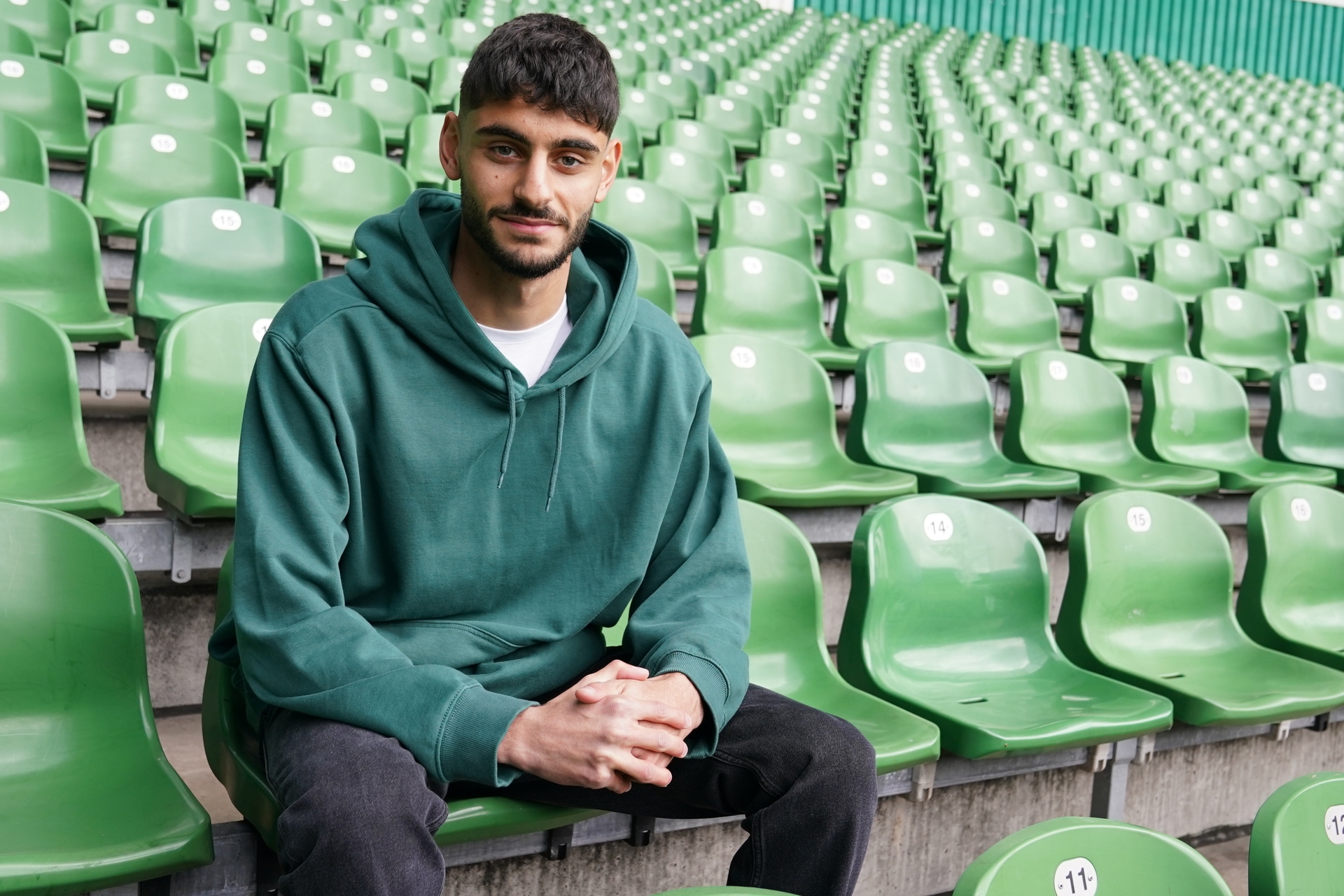 Eine Person in einem grünen Kapuzenpulli sitzt in einem fast leeren Stadion mit grünen Sitzen.