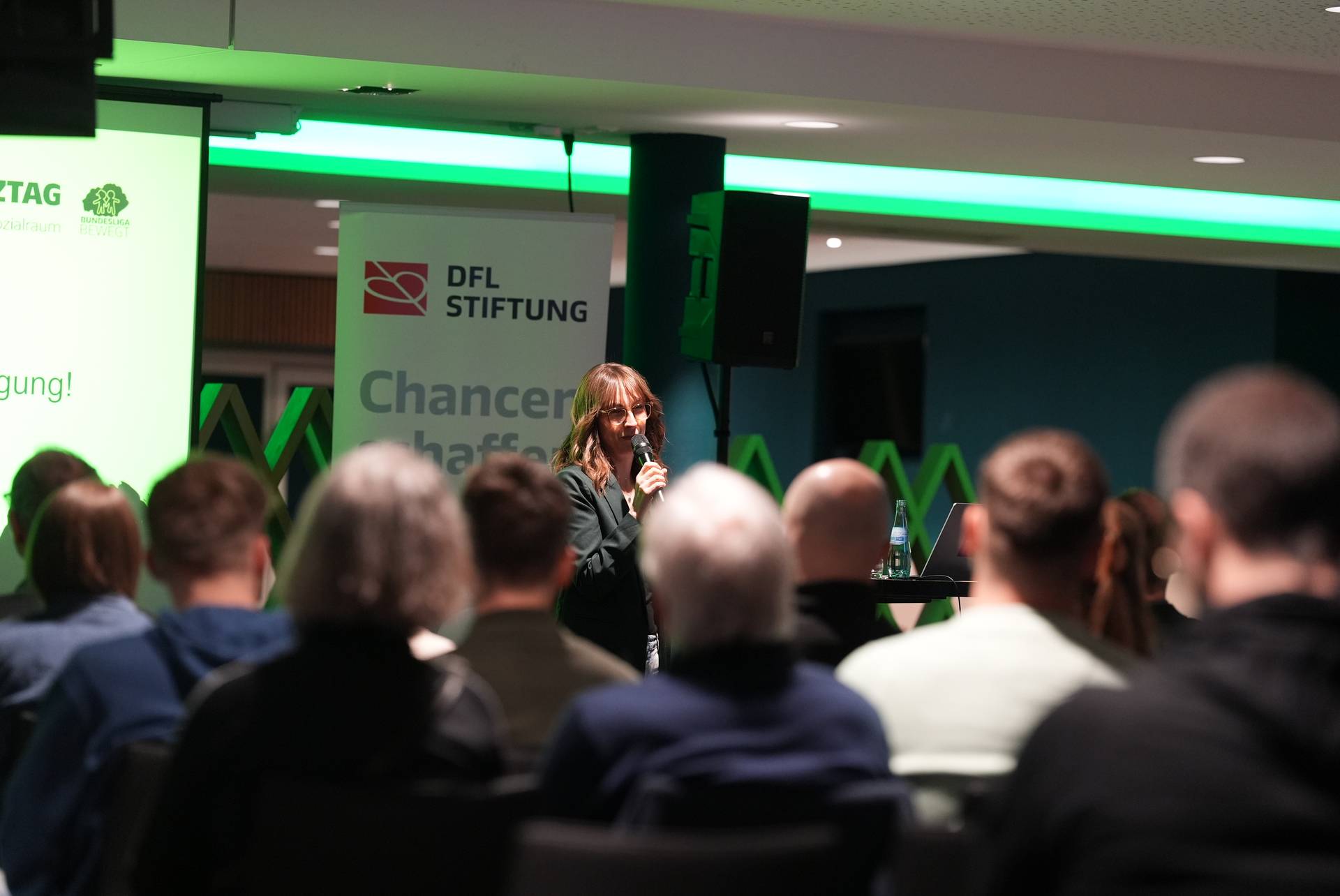 Eine Rednerin spricht bei einer Veranstaltung vor Publikum, neben ihr sind Banner mit den Aufschriften „DFL Stiftung” und „Chancen schaffen” zu sehen. Im Vordergrund sitzen Menschen.