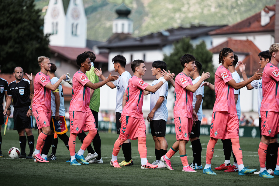 Zwei Fußballmannschaften in rosa-weißen Trikots geben sich auf dem Spielfeld die Hand, im Hintergrund sind Schiedsrichter und eine bergige Landschaft zu sehen.