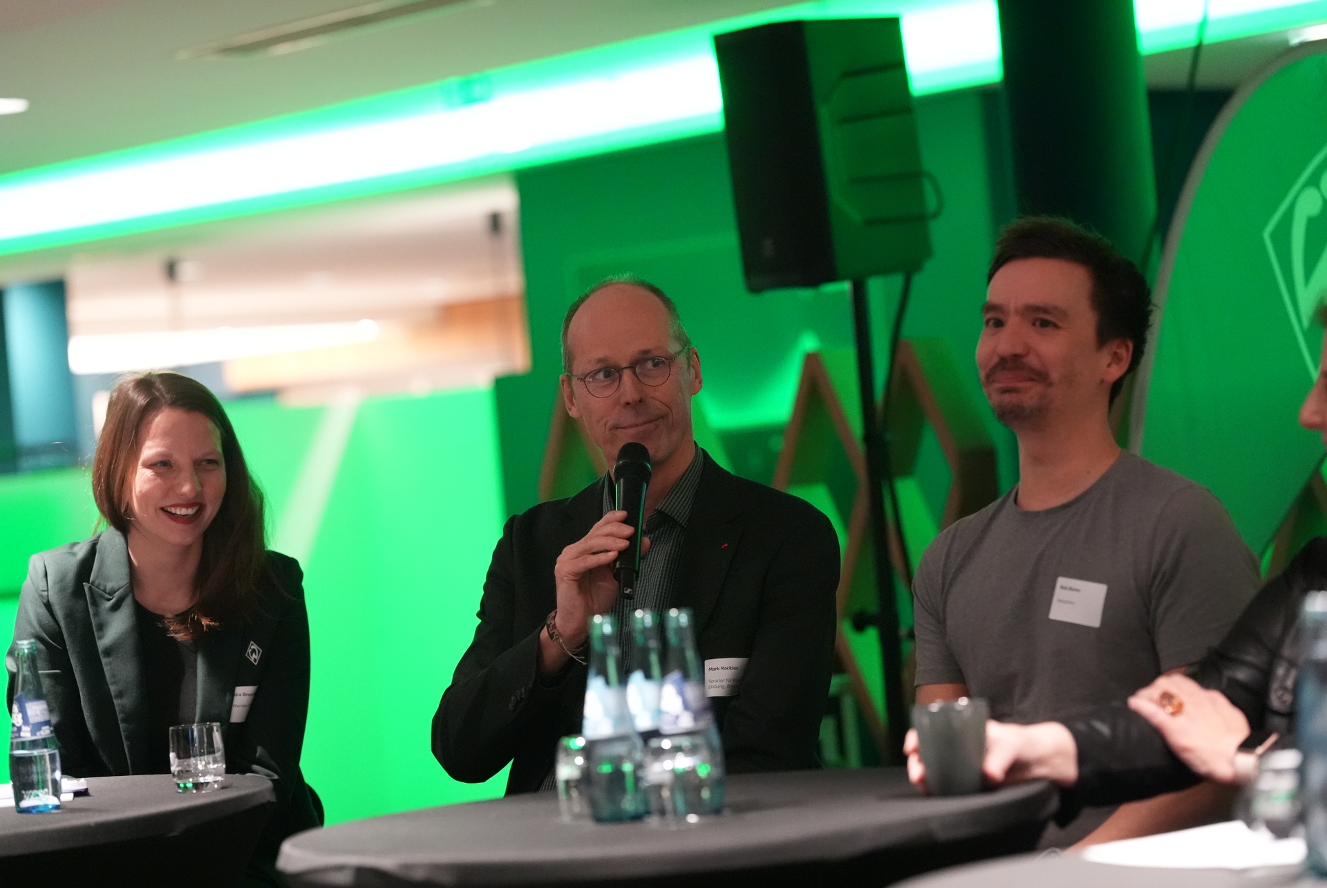 Drei Personen sitzen bei einer Podiumsdiskussion. Der Mann in der Mitte spricht mit einem Mikrofon, während die anderen lächelnd zuhören.