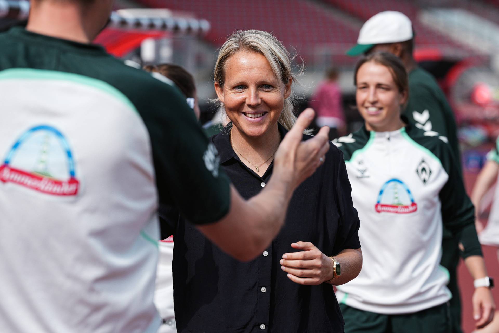 Eine lächelnde Frau in einem schwarzen Shirt steht zusammen mit Menschen in Sportkleidung auf einem Sportplatz und unterhält sich mit ihnen.