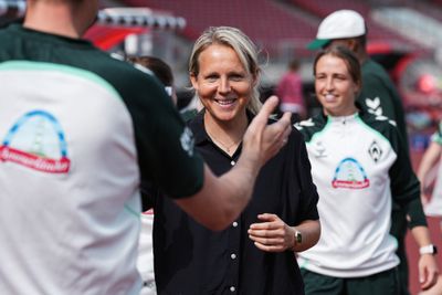 Eine lächelnde Frau in einem schwarzen Shirt steht zusammen mit Menschen in Sportkleidung auf einem Sportplatz und unterhält sich mit ihnen.