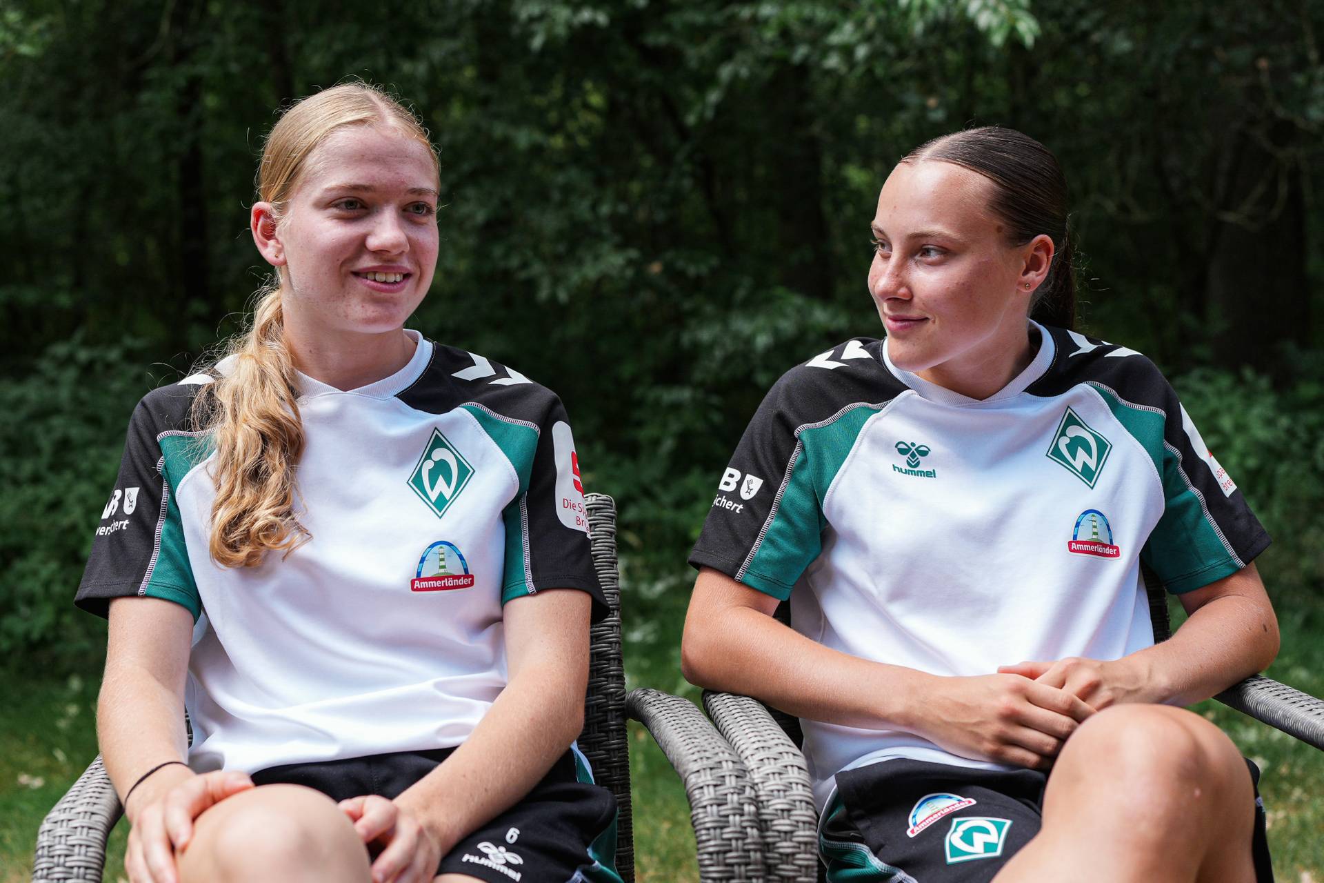 Zwei Frauen in grün-weißen Sporttrikots sitzen im Freien und schauen sich lächelnd an. Sie sind von Grün umgeben.