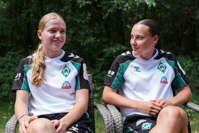 Zwei Frauen in grün-weißen Sporttrikots sitzen im Freien und schauen sich lächelnd an. Sie sind von Grün umgeben.