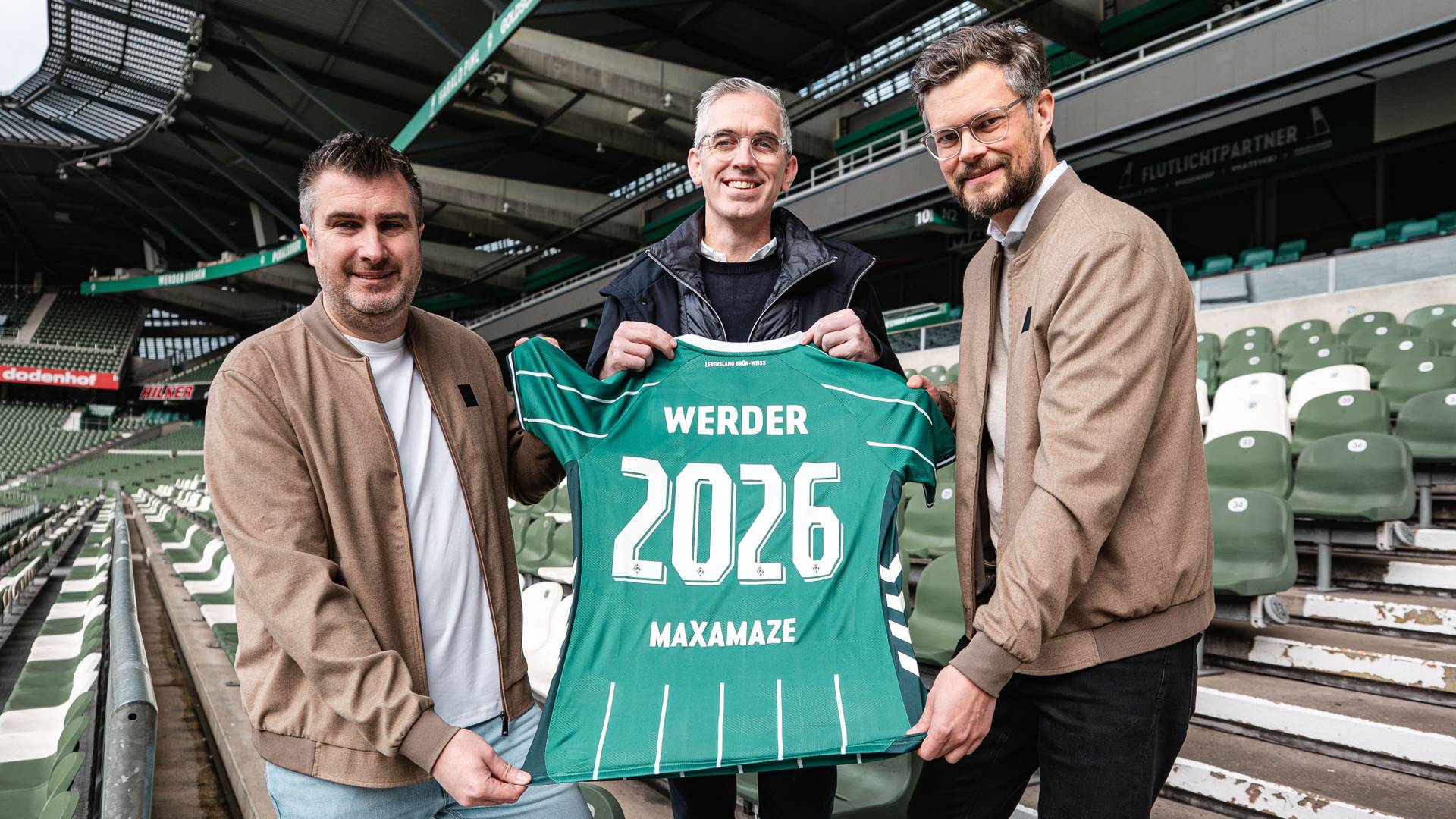 Drei Männer halten ein grünes Werder-Trikot mit der Aufschrift „MAXAMAZE 2026“ in den Händen und stehen in einem Stadion mit leeren Sitzplätzen.
