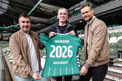 Drei Männer halten ein grünes Werder-Trikot mit der Aufschrift „MAXAMAZE 2026“ in den Händen und stehen in einem Stadion mit leeren Sitzplätzen.