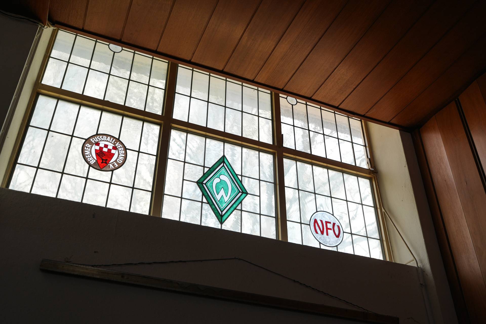 Buntglasfenster mit Sportlogos, bestehend aus einem roten Emblem, einem grünen rautenförmigen Logo und einem weißen Kreis mit der Aufschrift „NFV“ in roten Buchstaben.