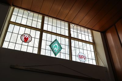 Buntglasfenster mit Sportlogos, bestehend aus einem roten Emblem, einem grünen rautenförmigen Logo und einem weißen Kreis mit der Aufschrift „NFV“ in roten Buchstaben.