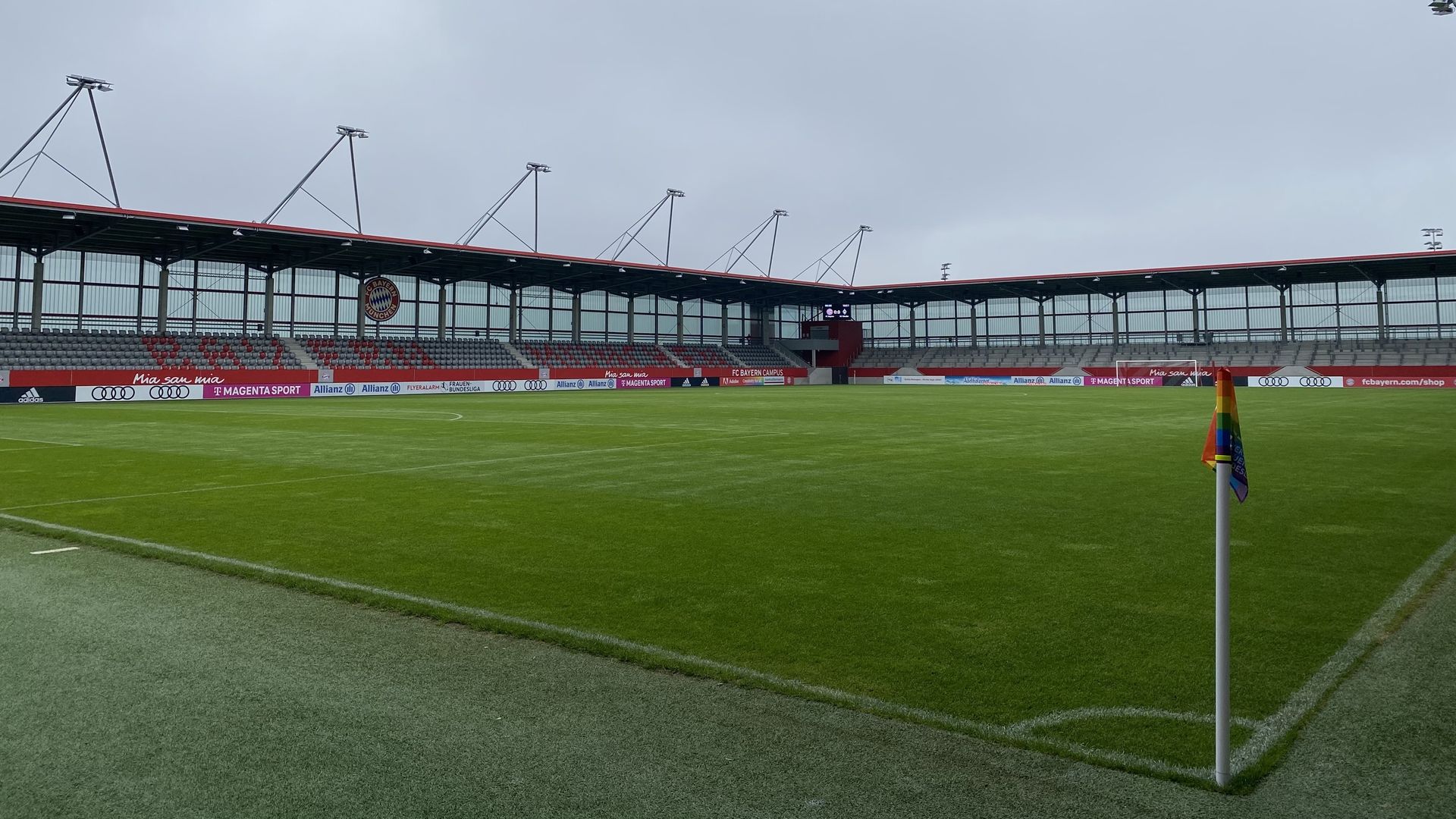 Leeres Fußballstadion mit grünem Rasen, roten und grauen Sitzplätzen und einer regenbogenfarbenen Eckfahne unter bewölktem Himmel.