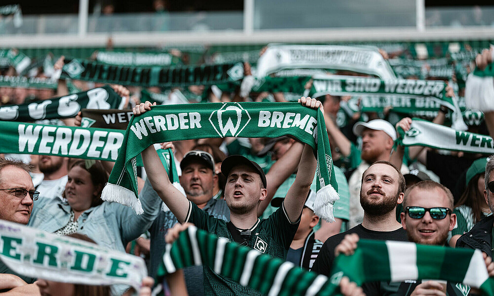 Menschen stehen auf einer Tribüne und halten grün-weiße Schals in die Höhe. 