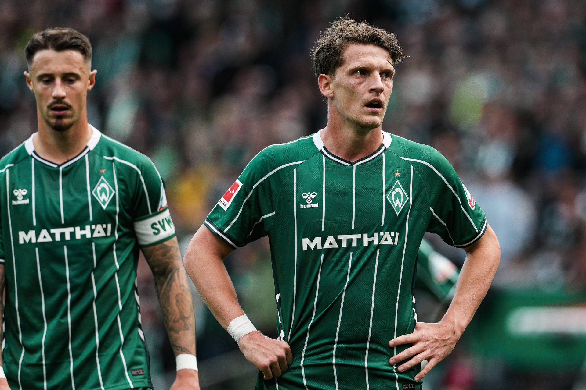 Zwei Fußballspieler in grünen Trikots mit weißen Streifen stehen auf dem Spielfeld. Der Spieler im Vordergrund hat die Hände in die Hüften gestützt und schaut konzentriert nach vorne, während der zweite Spieler links im Bild leicht unscharf wirkt. Im Hintergrund ist ein Stadion mit vielen Zuschauern zu sehen, die ebenfalls unscharf dargestellt sind.