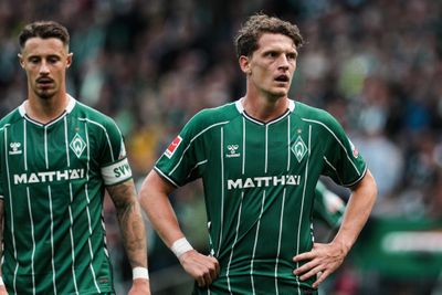 Zwei Fußballspieler in grünen Trikots mit weißen Streifen stehen auf dem Spielfeld. Der Spieler im Vordergrund hat die Hände in die Hüften gestützt und schaut konzentriert nach vorne, während der zweite Spieler links im Bild leicht unscharf wirkt. Im Hintergrund ist ein Stadion mit vielen Zuschauern zu sehen, die ebenfalls unscharf dargestellt sind.