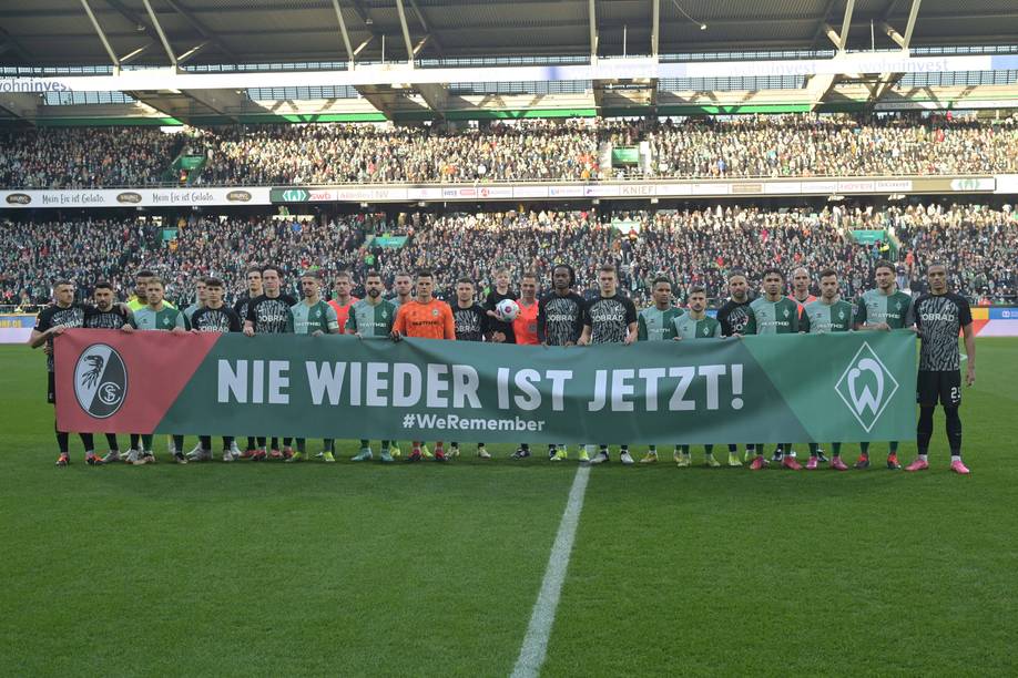 Fußballmannschaften stehen auf einem Spielfeld und halten ein Transparent mit der Aufschrift „Nie Wieder Ist Jetzt!“ hoch, im Hintergrund ist eine große Menschenmenge im Stadion zu sehen.