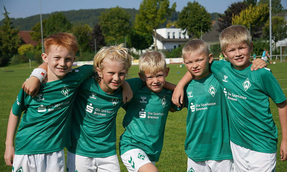 Kinder stehen Arm in Arm auf dem Fußballplatz und lächeln in die Kamera.