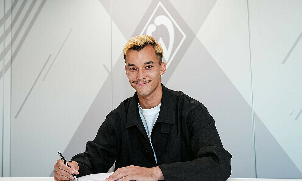 Mio Backhaus unterschreibt seinen Vertrag und schaut dabei in die Kamera. 
