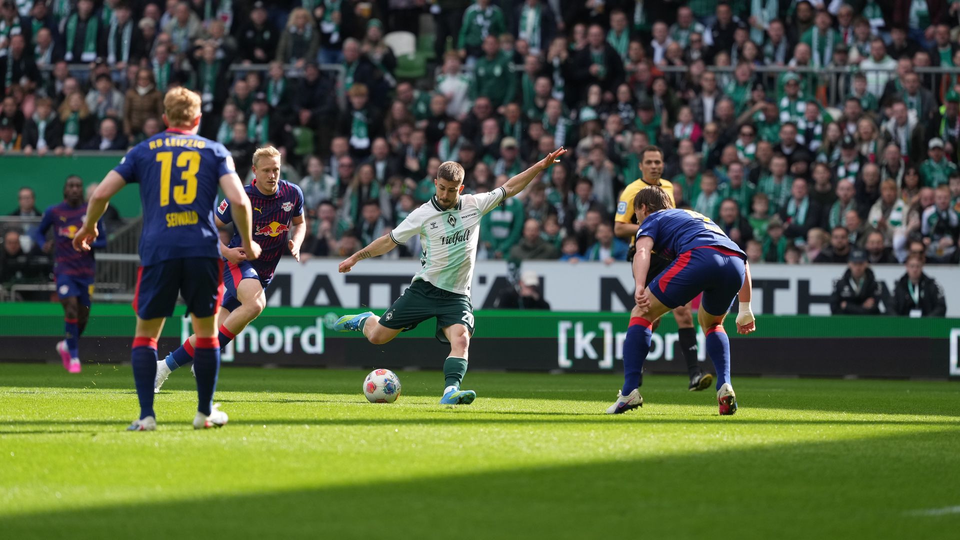A soccer player in a white and green kit prepares to kick the ball amidst opposing players on a sunny field, with spectators in the background.