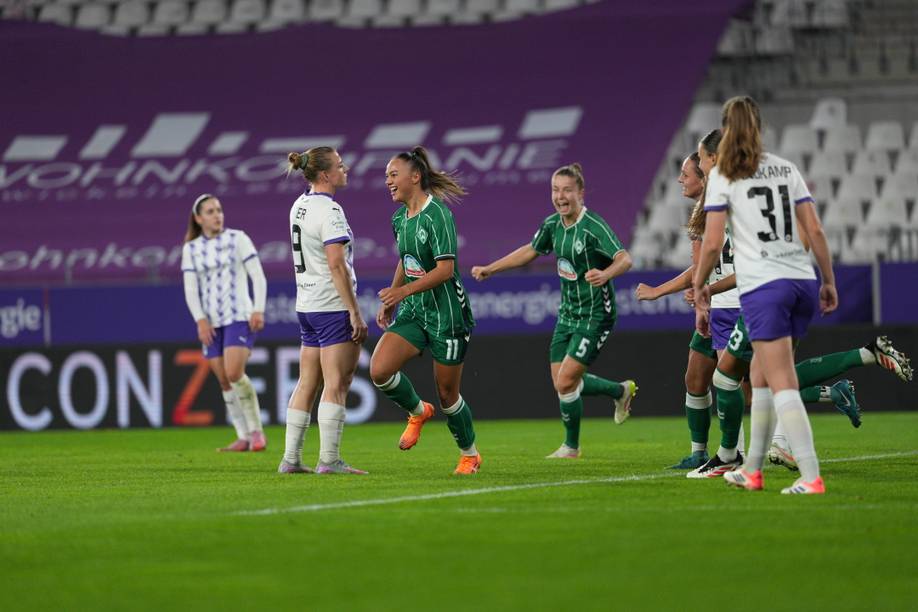 Eine Fußballspielerin jubelt nach einem Tor. Sie trägt ein grünes Trikot mit Werder-Raute. Um sie herum stehen Spielerin in weißen Trikots und sind enttäuscht.