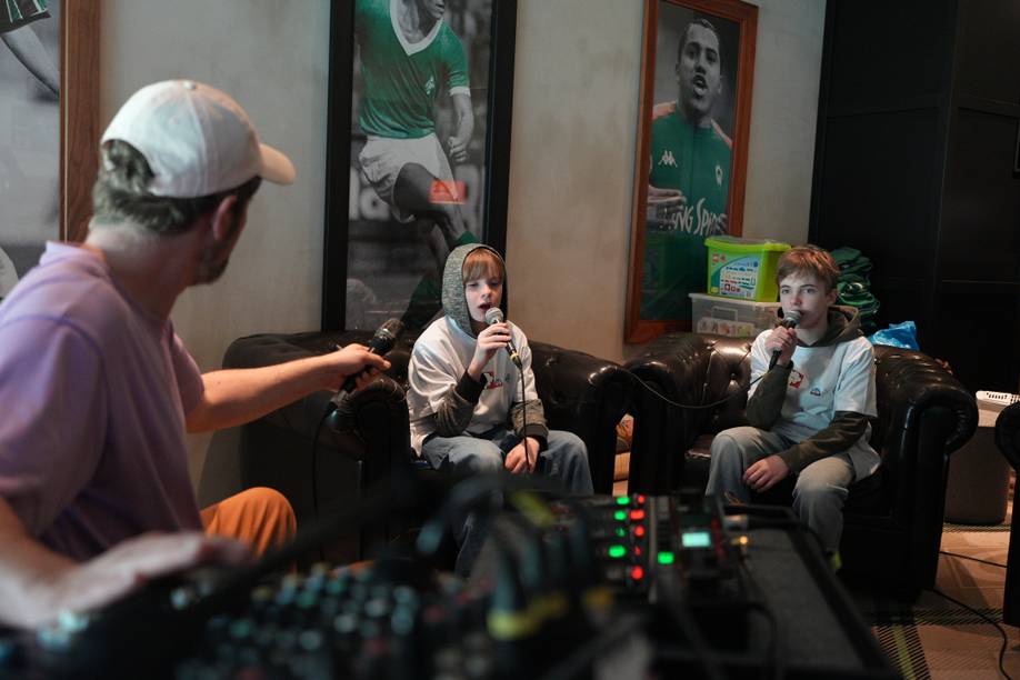 Drei Personen sitzen in einem Raum mit Sportpostern und sprechen in Mikrofone; im Vordergrund ist ein Mischpult zu sehen.