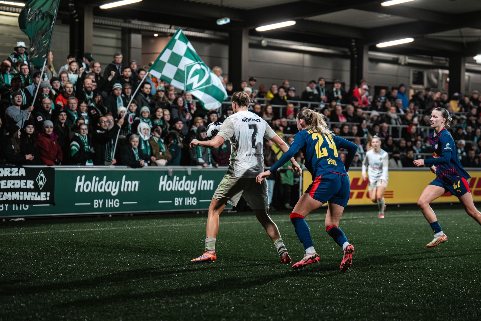 Larissa Mühlhaus im Zweikampf mit einer Spielerin von Leipzig, im Hintergrund sieht man die Werder-Fans im Gäste-Block.