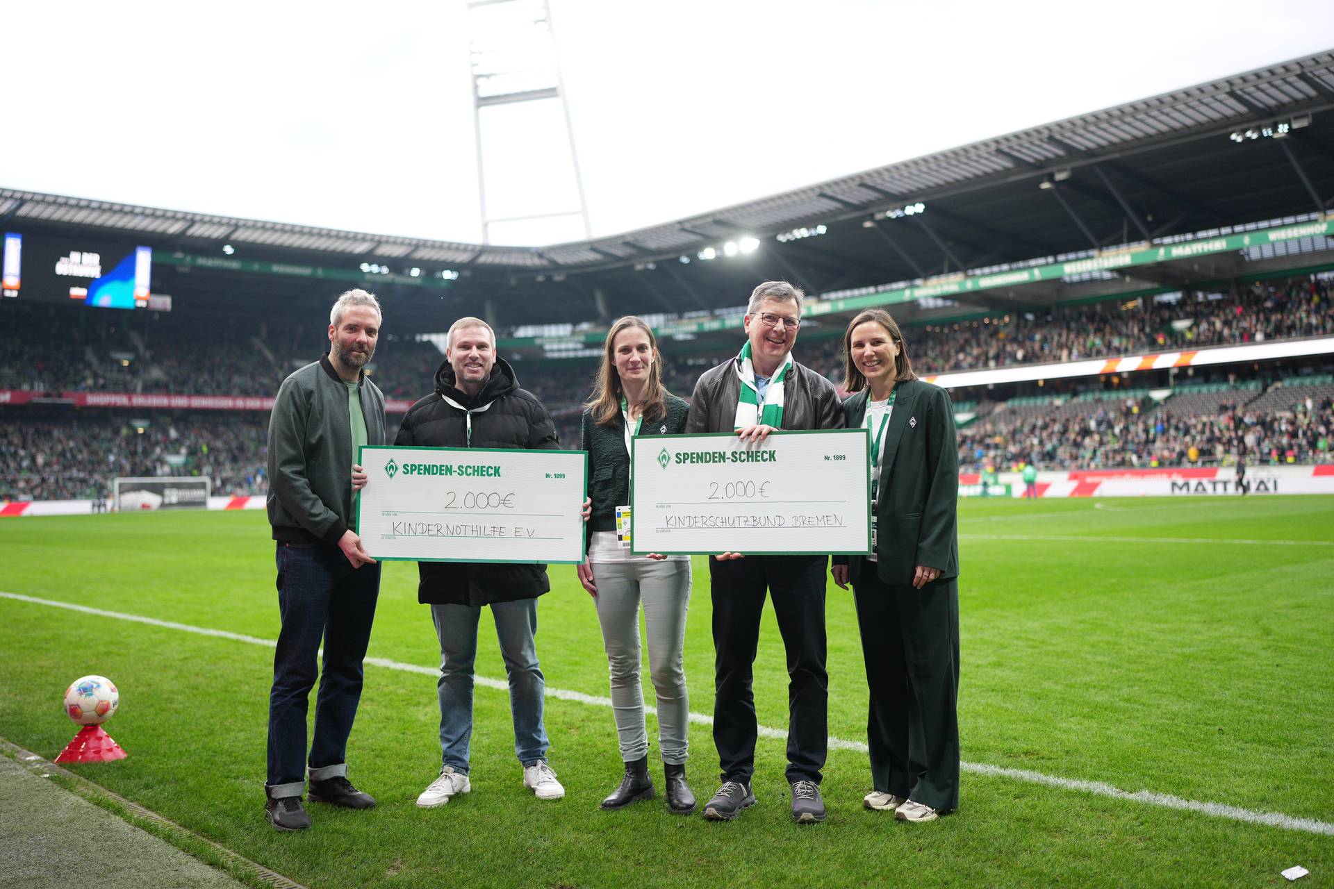 Fünf Personen stehen auf einem Fußballfeld und halten zwei große Spendenschecks in den Händen, im Hintergrund sind ein Stadion und eine Menschenmenge zu sehen.